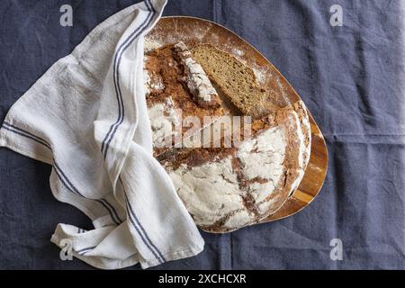 Photographie de table d'un pain de seigle frais cuit coupant en deux et tranchant sur une planche à découper en bois d'acacia recouverte d'une serviette de cuisine blanche sur un b foncé Banque D'Images