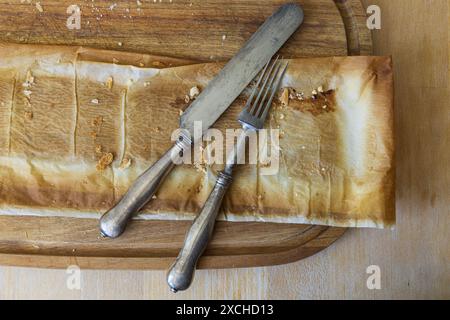 Photographie rapprochée sur table d'une planche à découper en bois d'acacia avec du papier sulfurisé laissé sur la tarte et fourchette vintage et couteau sur une table en bois Banque D'Images