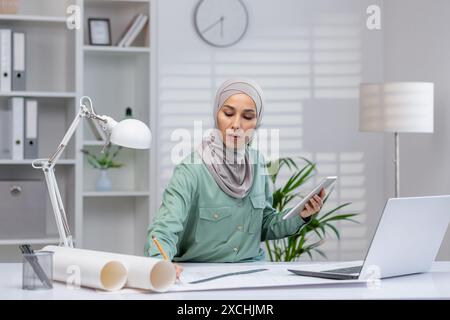 Femme architecte en hijab travaillant sur un projet utilisant une tablette et un ordinateur portable dans un cadre de bureau contemporain. Banque D'Images