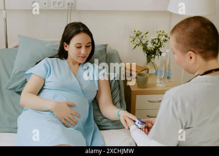 Au-dessus de l'épaule tir d'une femme enceinte allongée sur le lit et souriant légèrement pendant que le médecin vérifie sa saturation Banque D'Images