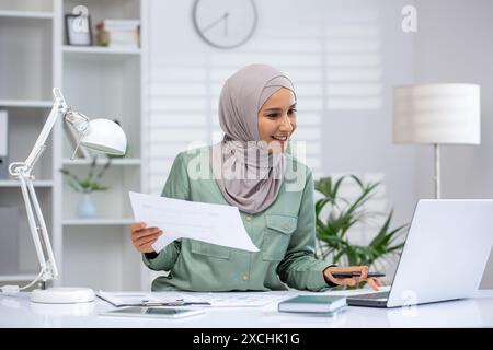 Femme d'affaires confiante dans le hijab travaillant au bureau à l'aide d'un ordinateur portable et de documents dans un bureau moderne. Cadre de travail professionnel au design contemporain et à la lumière naturelle. Banque D'Images