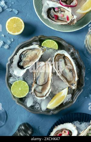 Huîtres sur un plat avec de la glace, avec du citron et du citron vert, plan de pose au-dessus plan sur un fond d'ardoise bleu Banque D'Images