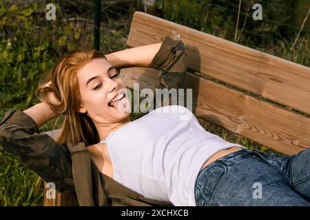 Jeune femme insouciante allongée sur un banc de parc en été, sortant sa langue de manière ludique, exsudant le bonheur et la détente sous le soleil chaud Banque D'Images