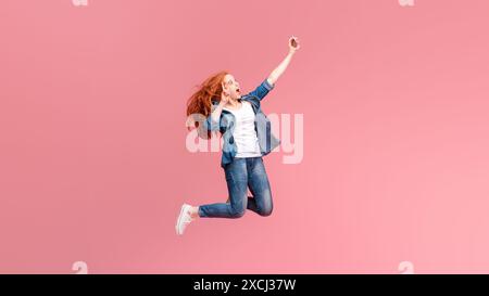 Joyeuse jeune femme aux cheveux rouges sautant haut sur un fond rose Banque D'Images