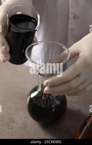 Femme versant du pétrole brut noir du bécher dans la fiole à la table grise, gros plan Banque D'Images