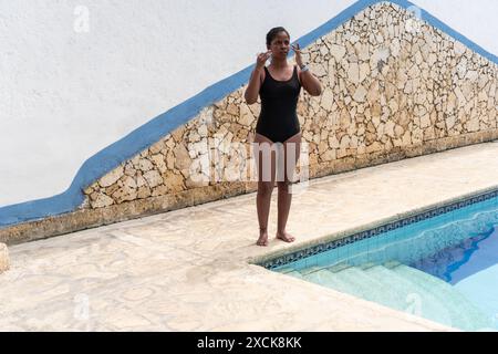 Une femme en maillot de bain noir se tient au bord d'une piscine, ajustant ses cheveux. Banque D'Images