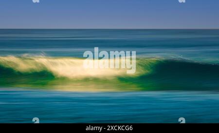 Une scène tranquille d'une douce vague se brisant sur une plage de sable alors que le soleil se couche, projetant une lueur chaude sur l'eau et le sable. L'eau est un turquoi vibrant Banque D'Images