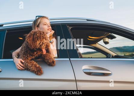 Portrait chien Maltipoo brun pelucheux avec une petite fille souriante regardant par une fenêtre de voiture ouverte alors qu'ils sont assis sur le siège arrière pendant le voyage en voiture. Fu Banque D'Images