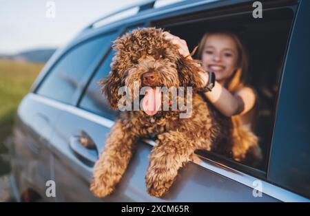 Portrait chien Maltipoo brun pelucheux avec une petite fille souriante regardant par une fenêtre de voiture ouverte alors qu'ils sont assis sur le siège arrière pendant le voyage en voiture. Fu Banque D'Images