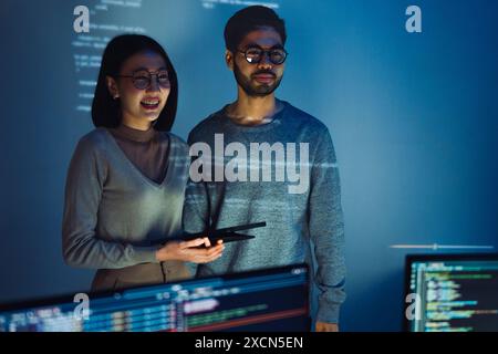 Asiatique deux développeurs de logiciels engagés dans une session de codage collaboratif, avec du code projeté sur le mur et plusieurs écrans d'ordinateur s'affichant Banque D'Images