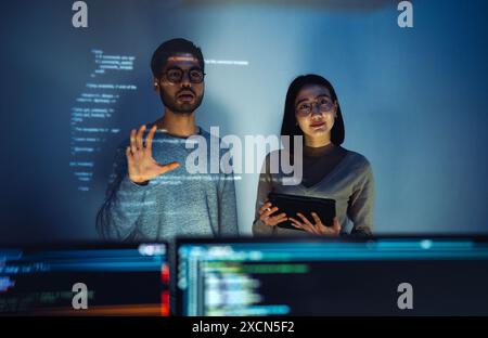 Asiatique deux développeurs de logiciels engagés dans une session de codage collaboratif, avec du code projeté sur le mur et plusieurs écrans d'ordinateur s'affichant Banque D'Images