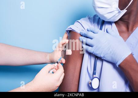 Paire de mains caucasiennes tenant seringue et aiguille donnant le vaccin à l'assistant médical noir. Médecin vaccinant un homme afro-américain et pose un bandage sur son bras. Banque D'Images