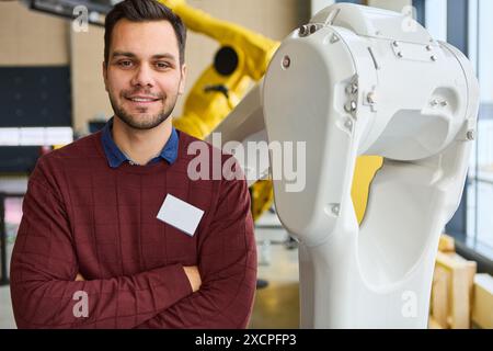 Ingénieur posant en toute confiance à côté d'un bras robotique blanc dans un environnement industriel moderne, mettant en avant la robotique et l'automatisation des processus. Banque D'Images