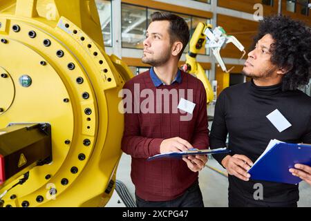 Deux ingénieurs examinent et programment des bras robotiques polyvalents dans une installation industrielle moderne, mettant en avant l'automatisation et la technologie. Banque D'Images