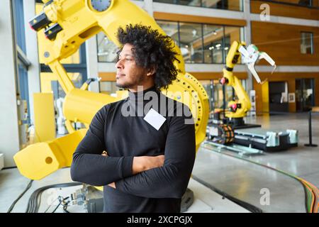 Jeune homme debout avec confiance dans un laboratoire de robotique avec des bras robotiques industriels jaunes en arrière-plan. Concept de robotique, d'automatisation et de moderne te Banque D'Images