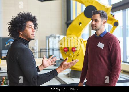 Deux hommes discutent du développement et de la programmation robotique dans un environnement industriel moderne avec un grand bras robotique jaune en arrière-plan. Banque D'Images