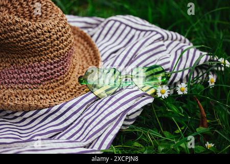 Lunettes de soleil en forme de coeur vert et chapeau de paille sur un tissu rayé sur une prairie verte fleurie avec des marguerites blanches. Nature morte estivale. Vacances d'été Banque D'Images