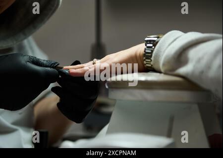 Gros plan d'une manucure pratiquant une manucure sur un client dans un salon de beauté. Le professionnel porte des gants noirs tout en façonnant les ongles. Banque D'Images