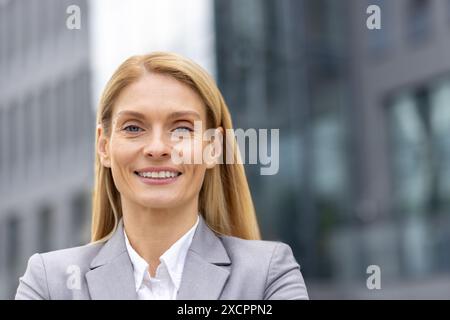 Portrait en gros plan d'une jeune femme d'affaires blonde dans un costume gris debout à l'extérieur d'un immeuble de bureaux, regardant et souriant à la caméra. Banque D'Images