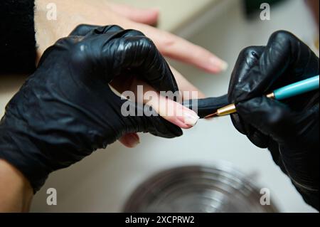 Gros plan d'une manucure portant des gants noirs, appliquant méticuleusement du vernis à ongles sur les ongles d'un client dans un salon de beauté. Parfait pour les concepts de soins, Banque D'Images