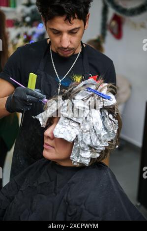 Coiffeur styliste travaillant sur les cheveux d'un client dans un salon de beauté, appliquant la teinture tout en utilisant des reflets de feuille. Services professionnels de coiffure. Banque D'Images