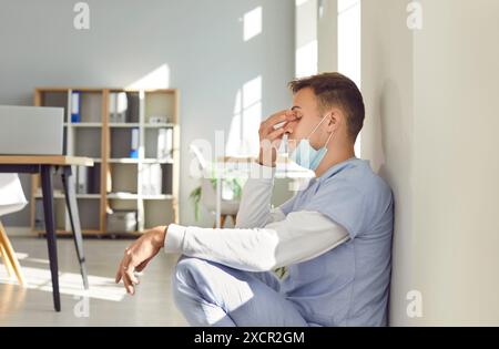 Médecin masculin fatigué stressé portant un uniforme bleu assis à l'hôpital souffrant de maux de tête. Banque D'Images