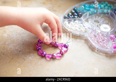 Fille d'âge préscolaire engagée dans l'activité d'art et d'artisanat, projet d'artisanat de bricolage, fabrication de bracelet personnalisé avec des perles colorées, motricité fine, promotion Banque D'Images