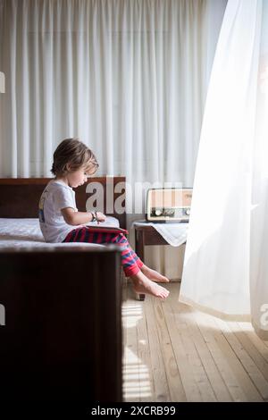 Un jeune garçon est assis sur un lit, lisant un livre. Le soleil brille brillamment à travers une fenêtre, projetant une lueur chaude sur la pièce. Banque D'Images