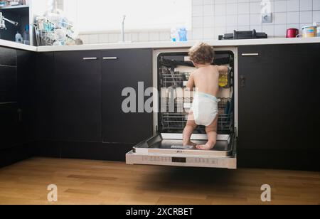 Un jeune enfant dans une couche se tient à l'intérieur d'un lave-vaisselle ouvert, explorant curieusement les étagères et les étagères. Banque D'Images