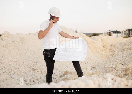 Ingénieur civil professionnel en casque de protection parle avec l'entrepreneur sur smartphone, discute des plans d'architecture, vérifiant le site de construction Banque D'Images