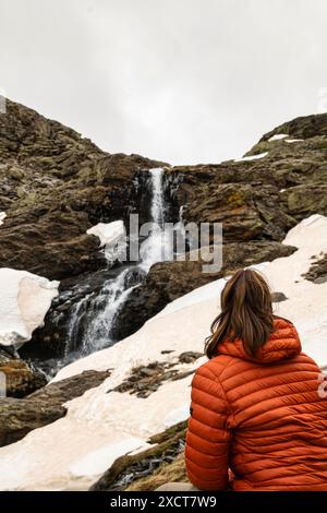 Fille assise sur son dos avec un plumeau orange et de longs cheveux regardant une cascade parmi les rochers et la neige dans le parc national de sierra nevada. Banque D'Images