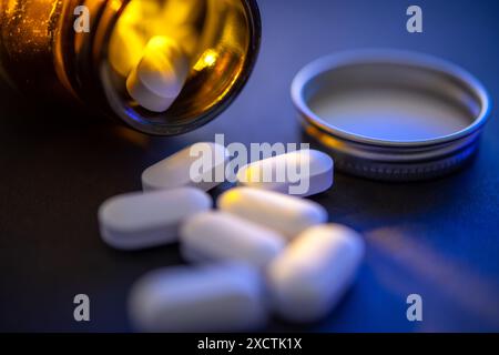 Gros plan de pilules blanches tombant d'une bouteille de médicament en verre ambré sur fond bleu sombre, mettant en évidence les soins pharmaceutiques et Banque D'Images