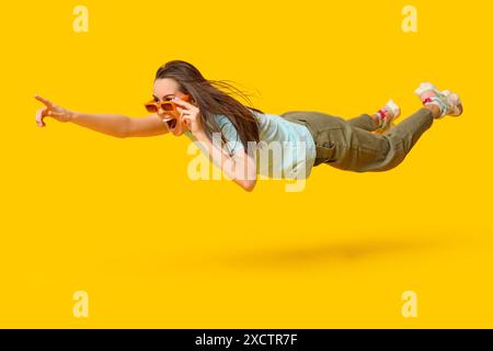 Surprise jeune femme dans des lunettes de soleil volant et pointant vers quelque chose sur fond jaune Banque D'Images