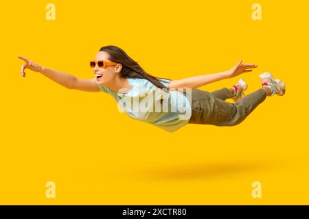 Surprise jeune femme dans des lunettes de soleil volant et pointant vers quelque chose sur fond jaune Banque D'Images