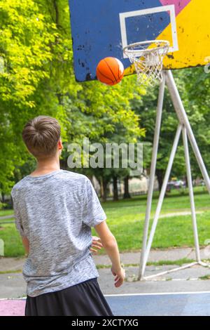 Un adolescent portant une chemise grise et un short noir joue au basket-ball sur un terrain extérieur. Le basket est en plein air, se dirigeant vers le cerceau, Banque D'Images