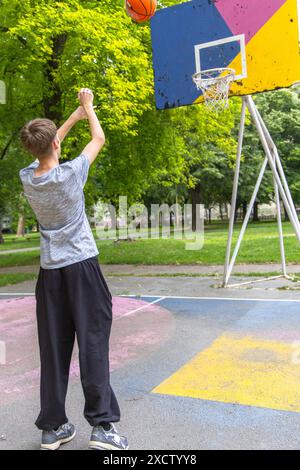 Un jeune homme vêtu d'un t-shirt gris et d'un pantalon noir se prépare à tirer sur un ballon de basket. Il est sur un terrain extérieur avec un panneau coloré et un panier Banque D'Images