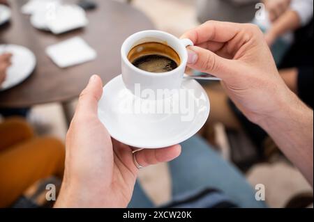 Gros plan des mains tenant une tasse blanche et une soucoupe avec du café expresso fraîchement préparé. La scène représente une pause café décontractée, la détente et le plaisir Banque D'Images