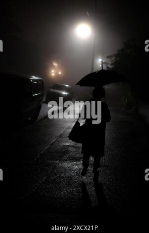 Silhouette d'une personne tenant un parapluie marchant à côté d'une route sombre sur une nuit brumeuse à Katoomba, des voitures garées et un éclairage de rue en avant Banque D'Images