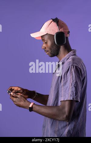 Homme afro-américain portant des écouteurs et utilisant un smartphone, vu de côté sur un fond violet. Idéal pour le contenu sur la technologie, Banque D'Images