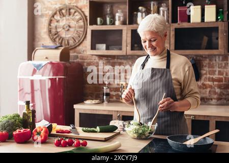 Femme senior mélangeant la salade dans la cuisine moderne avec réfrigérateur rouge Banque D'Images