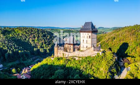 Le château de Karlstejn se dresse au sommet d'une colline, entouré de forêts vertes denses sous un ciel bleu clair, avec un village pittoresque niché dans la vallée en contrebas. République tchèque Banque D'Images