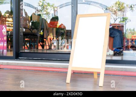 Un tableau blanc se tient debout sur un stand en bois à l'extérieur d'un restaurant. Le plateau est vide et le restaurant est visible à travers la porte vitrée Banque D'Images