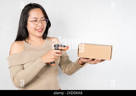 Une femme tient un scanner de code-barres et le pointe vers une boîte. Elle est souriante et elle est heureuse sur un fond blanc, un espace vide. Banque D'Images
