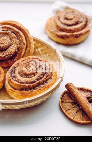 Petits pains de cannelle faits maison fraîchement cuits avec du sucre sur le dessus dans le panier dans la cuisine maison sur la table blanche, bâton de cannelle pour la décoration. Mise au point sélective. Banque D'Images