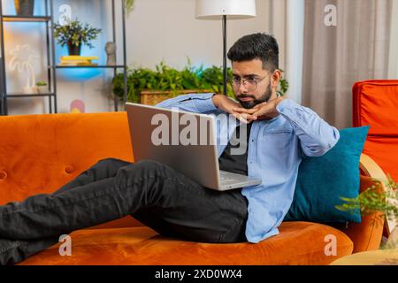 Bouleversé jeune homme indien assis sur le canapé à la maison à l'aide d'un ordinateur portable ayant l'anxiété et le stress. Un arabe choqué et barbu réagissant à la perte, de mauvaises nouvelles sur netbook dans l'appartement du salon. Virus informatique. Banque D'Images