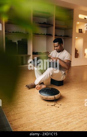 Plan vertical d'un homme africain jouant sur un instrument de musique moderne main pan, Vadjraghanta, tambour de langue en métal assis jambes croisées sur le sol à la maison. Banque D'Images