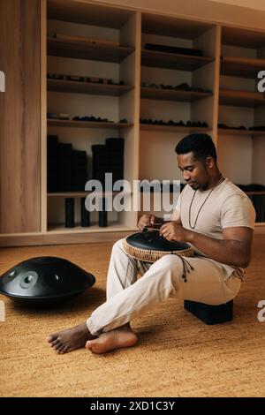 Plan vertical d'un homme africain jouant de la musique ethnique sur un tambour de réservoir en utilisant des baguettes de tambour assis sur le sol dans la pose de lotus. Serein noir pratiquant natif Banque D'Images