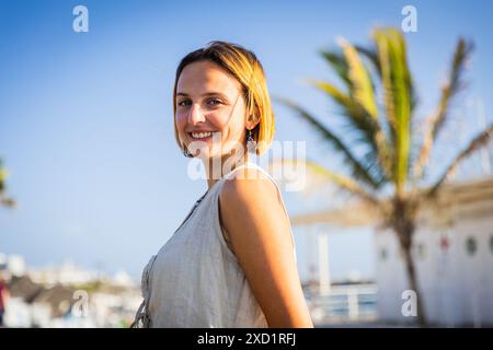 Une femme est souriante et debout devant un palmier, une journée ensoleillée créant une atmosphère positive. Banque D'Images