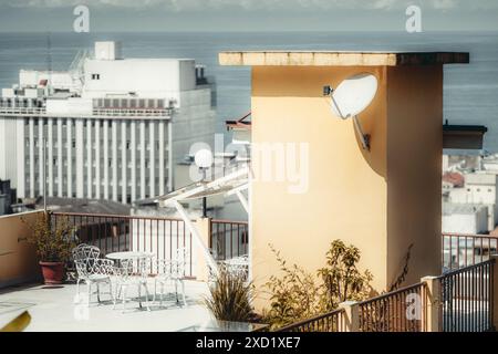 Terrasse sur le toit avec mobilier en fer forgé blanc, antenne parabolique et plantes en pot, avec vue sur l'océan et édifices urbains en toile de fond. L'image CA Banque D'Images