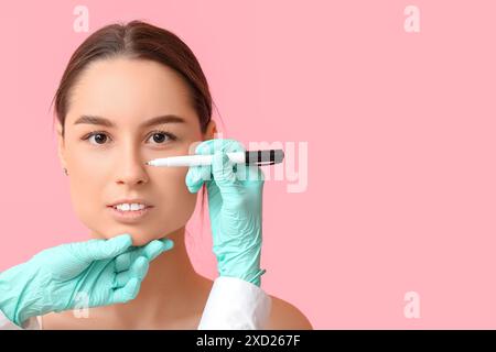 Docteur en gants de protection dessinant des marques sur le nez de la femme pour la chirurgie plastique sur fond rose Banque D'Images